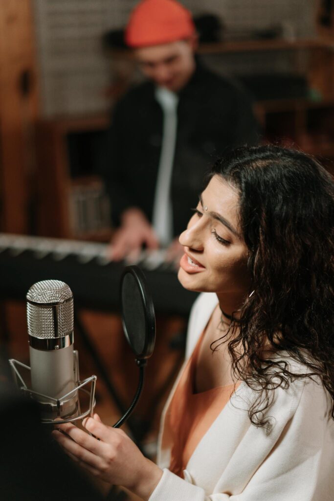 Woman singing into microphone with musician at keyboard in a recording studio.